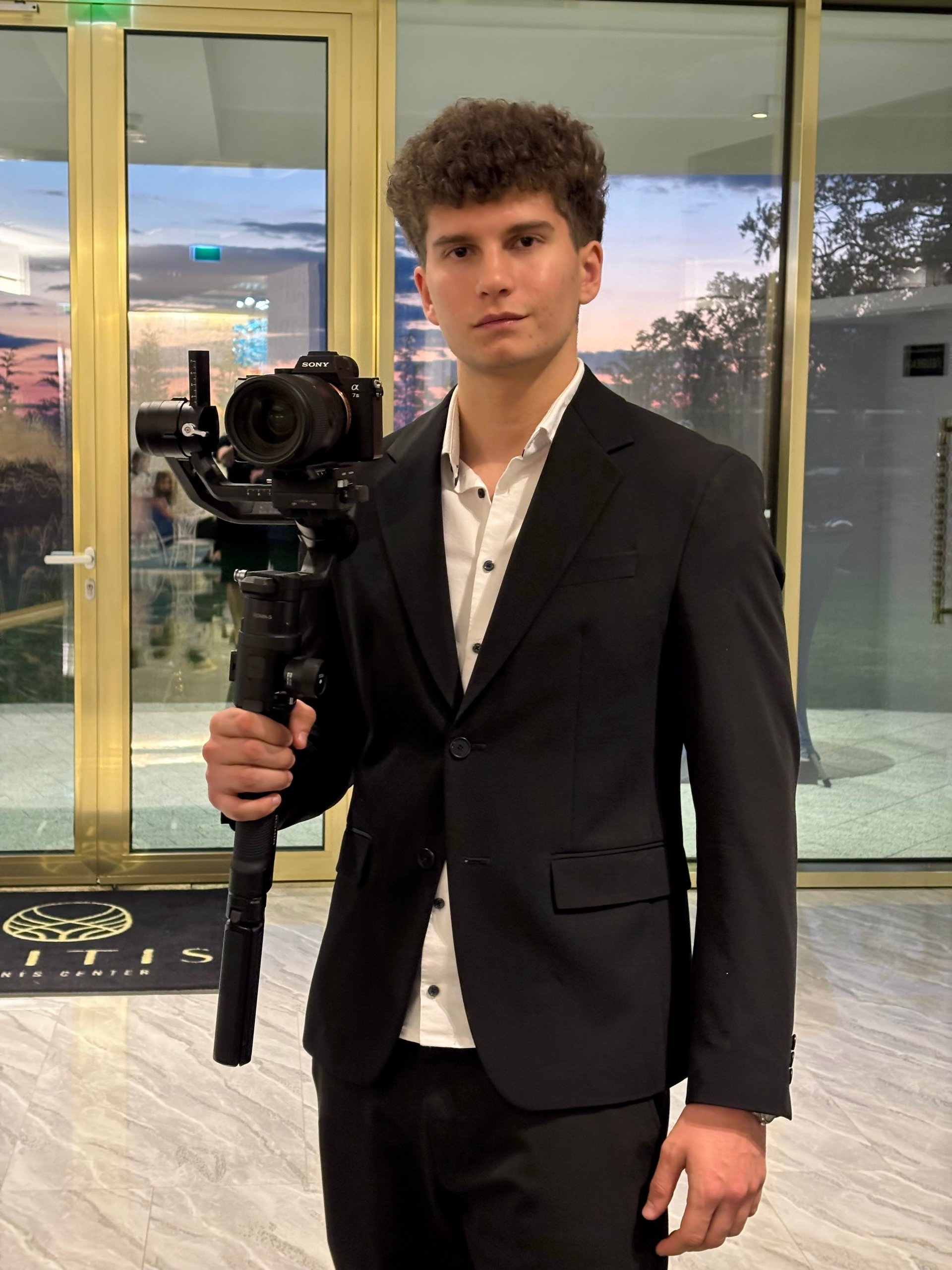 Young man in black suit holding professional camera on gimbal stabilizer indoors with windows in background